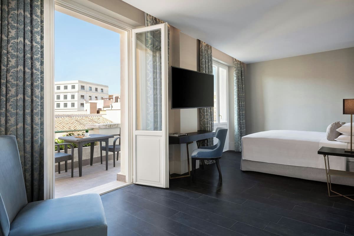 A hotel room with a bed, a desk with a chair, a TV, and a balcony with a table and chairs.