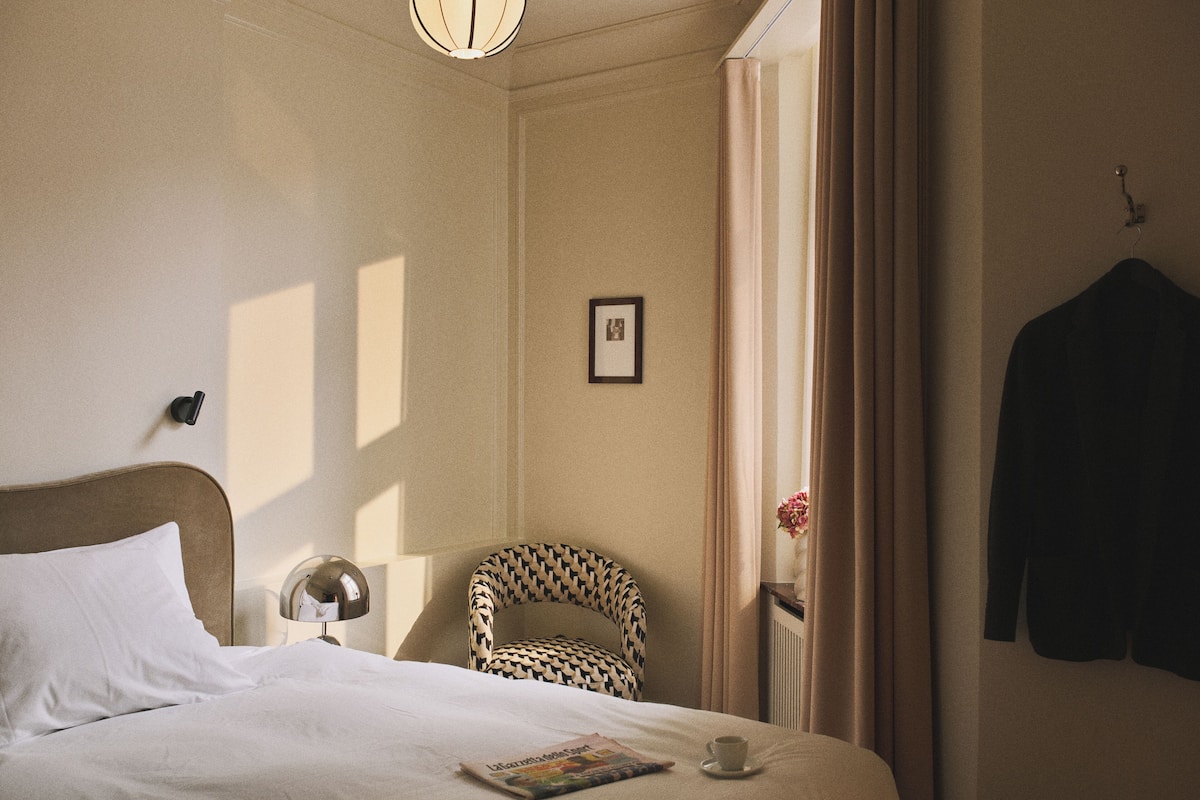 A hotel room with a bed, a chair, a lamp, a coat hanger, and a framed picture on the wall.