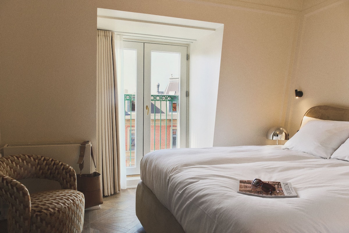 A hotel room with a bed, a chair, a window with curtains, and a view of buildings outside.