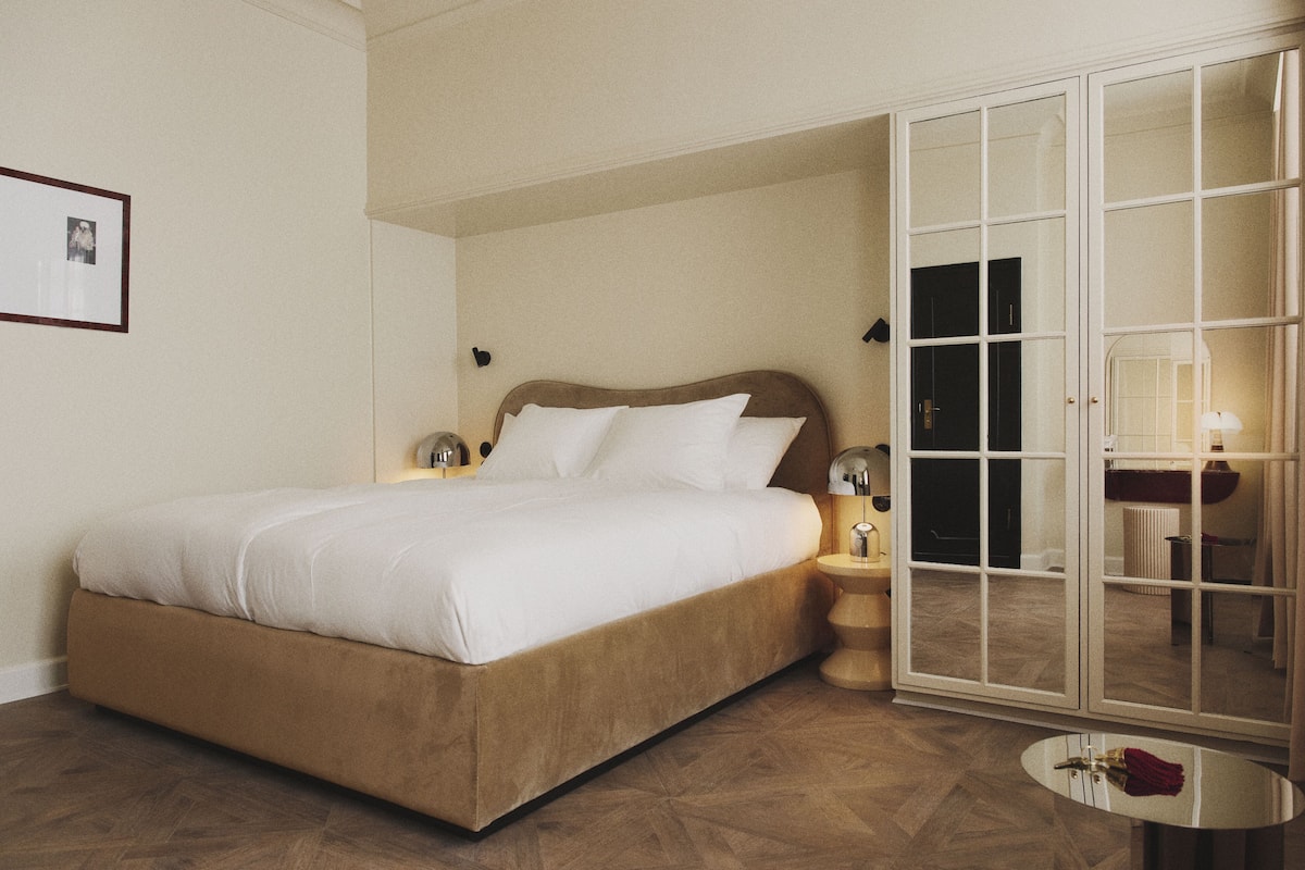 A neatly made bed with a headboard, a bedside table with a lamp, and a large mirror with a door.