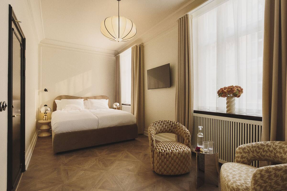 A hotel room with a large bed, a chair, a small table with glasses, a vase with flowers, and a TV on the wall.