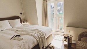 A hotel room with a large bed, a chair, a small table with a glass of wine, and a view of buildings through a window. - Hotel Bella Grande (Copenhagen)