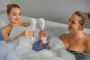 Dos personas en una bañera llena de burbujas, sosteniendo copas con un logotipo.