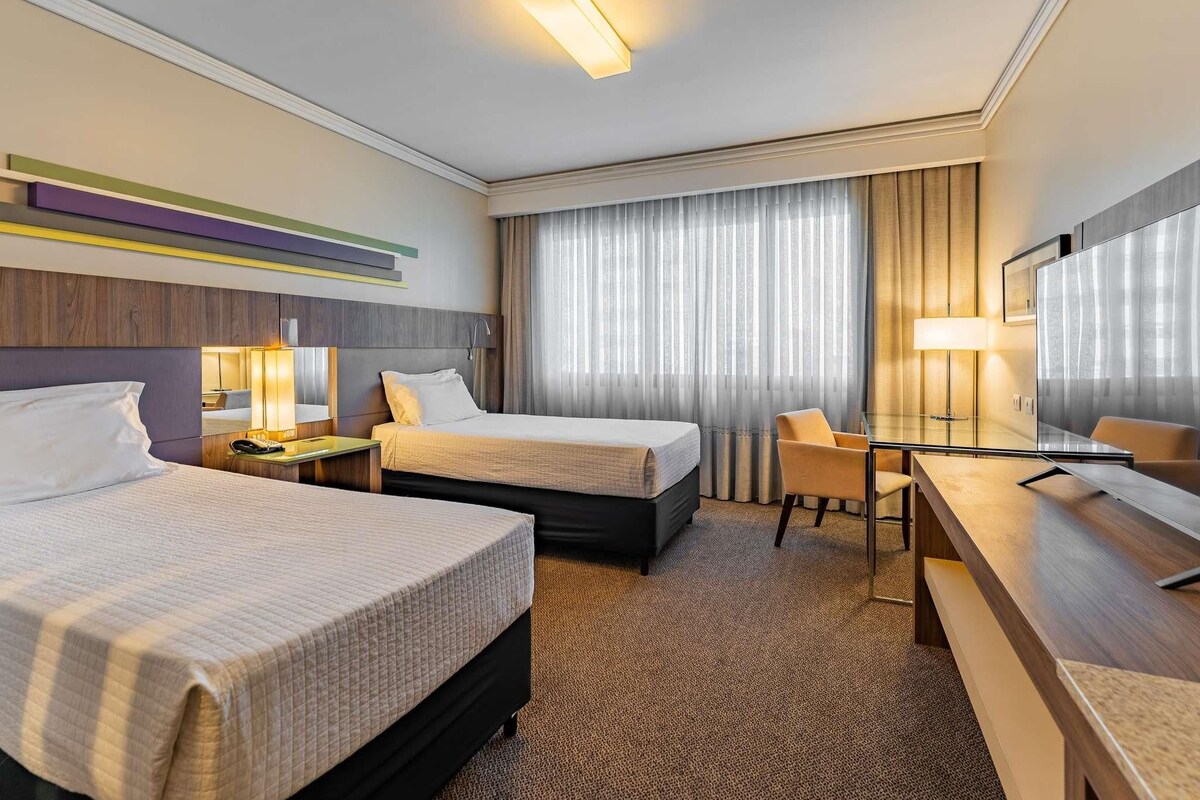 A hotel room with two beds, a desk, a chair, a bedside table, and a large window with curtains.