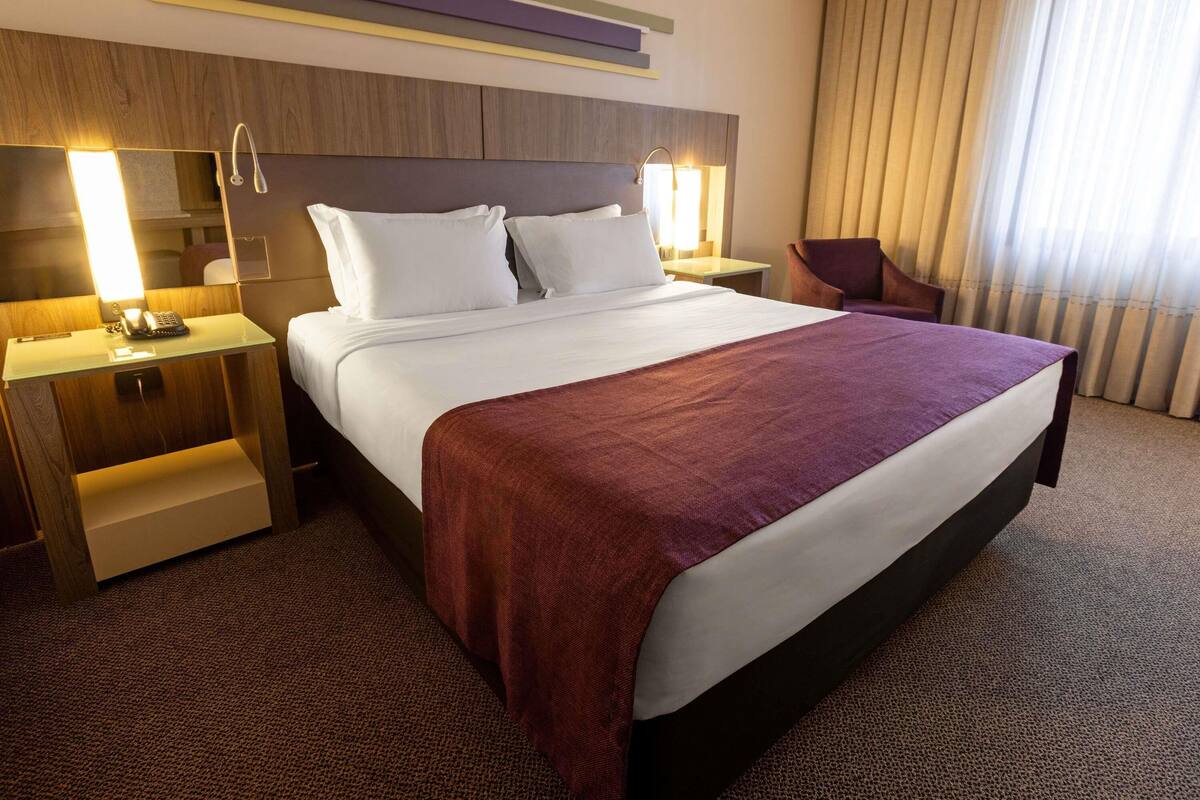 A hotel room with a large bed, bedside tables, a desk, a chair, and a window with curtains.