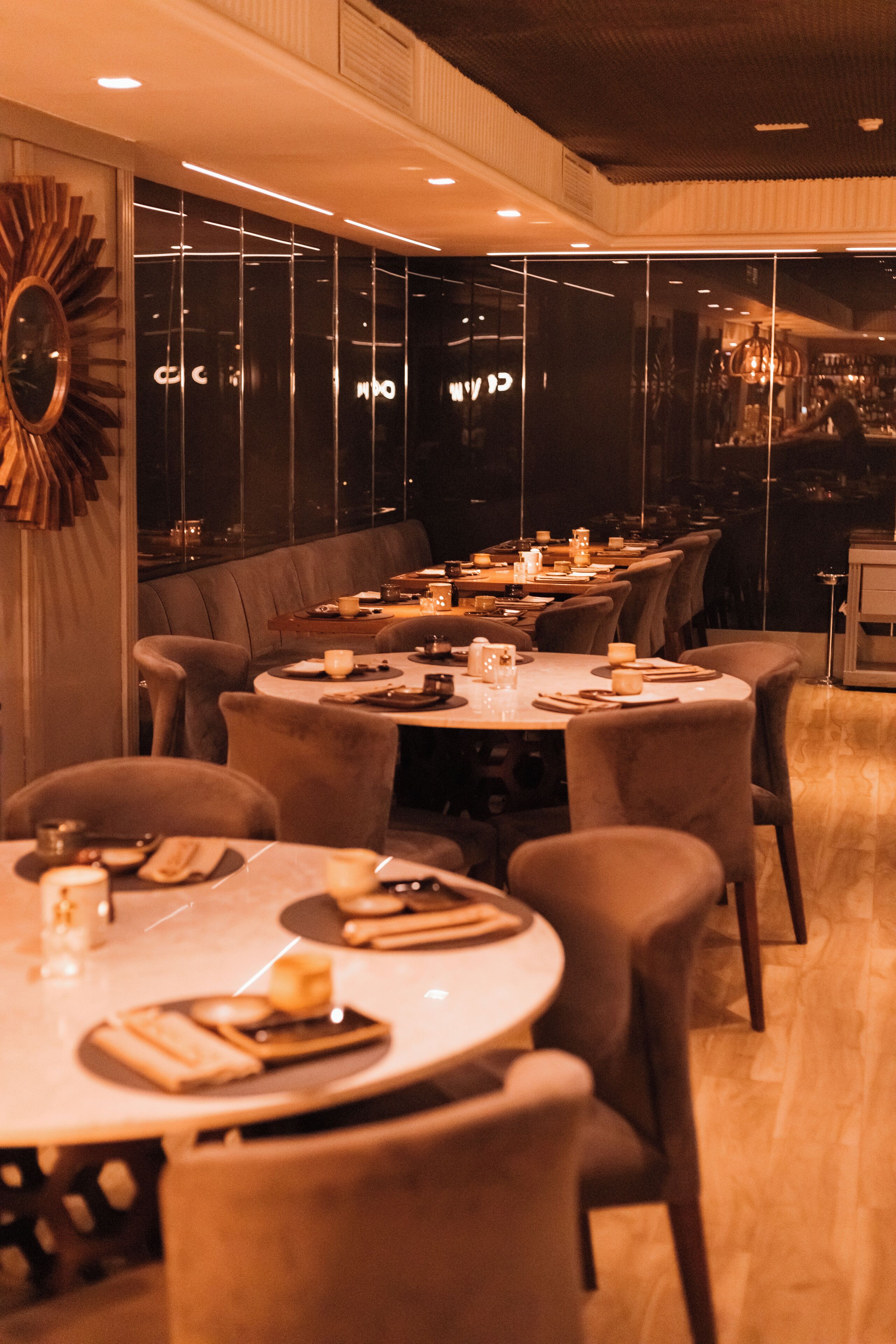 Un restaurante con mesas redondas y sillas acolchadas, un espejo grande en la pared y vistas a través de los ventanales.