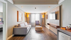 A hotel room with a sofa, ottoman, armchair, TV, and a dining area with a refrigerator. - Grand Mercure Curitiba Rayon (Curitiba)