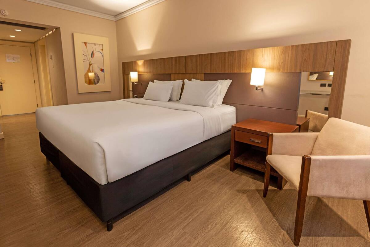 A hotel room with a large bed, bedside tables, a chair, and a wooden headboard.
