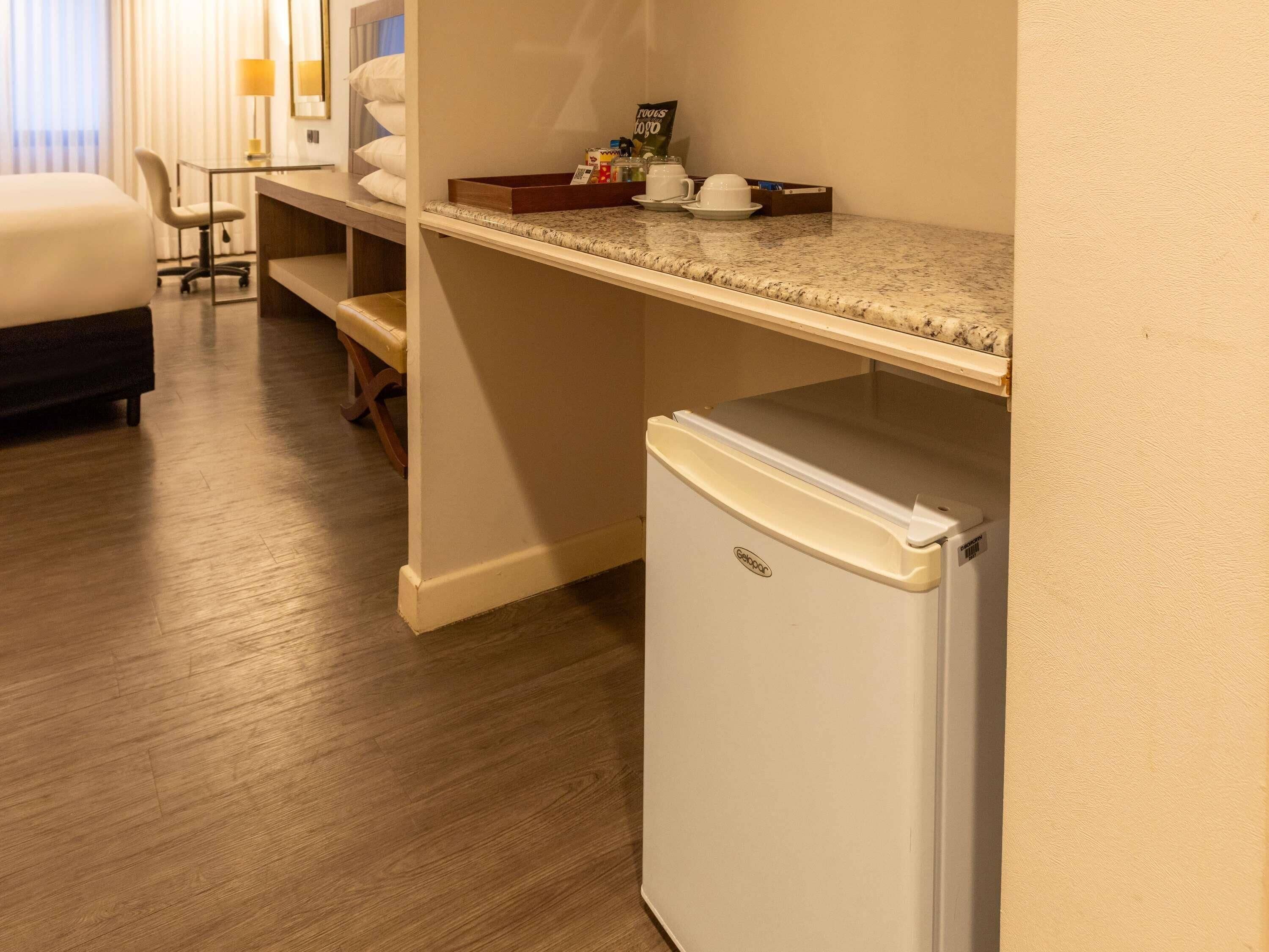 A compact hotel room with a bed, a desk, a chair, a small refrigerator, and a window with curtains.