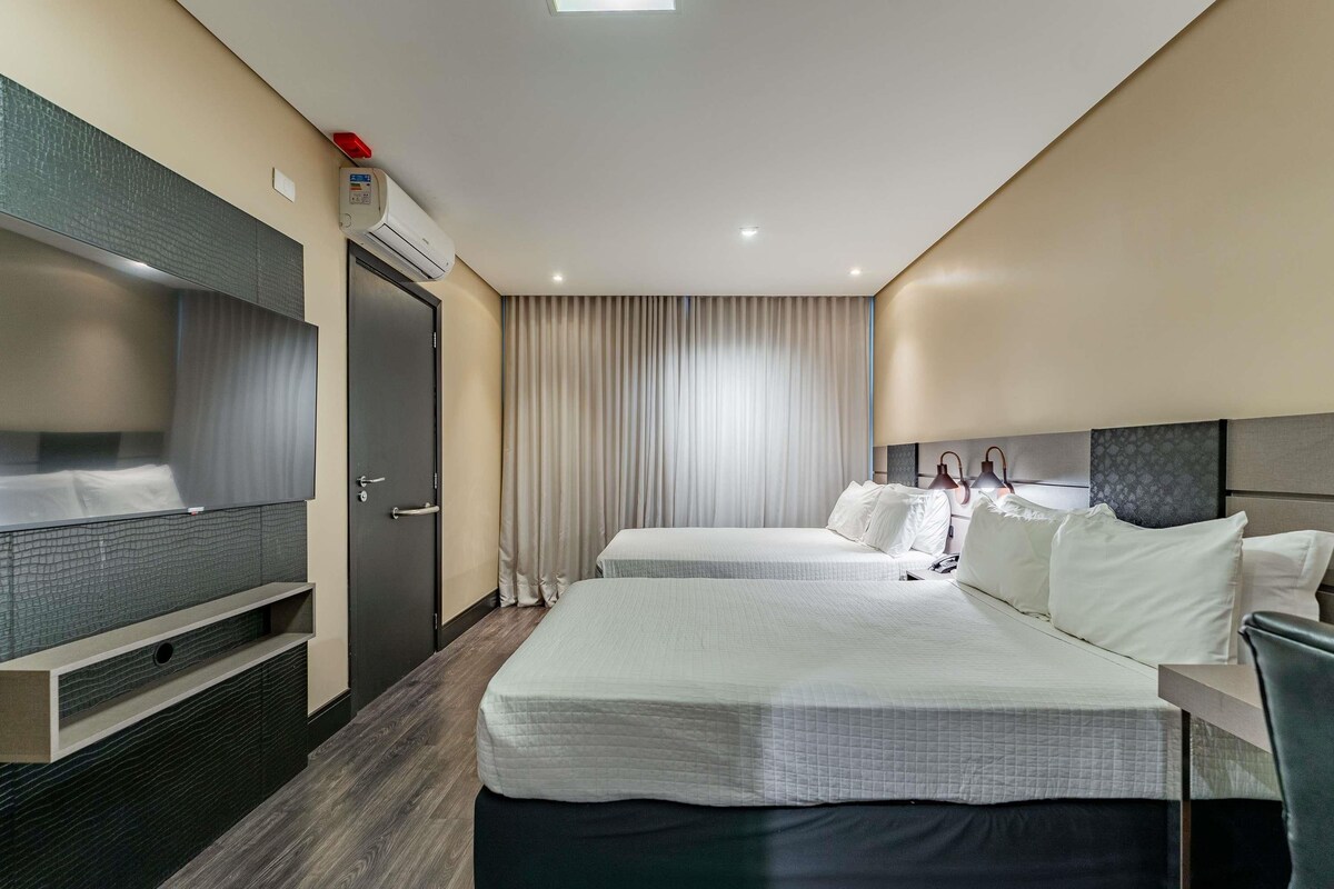 A hotel room with two beds, a TV, a desk, and a window with curtains.