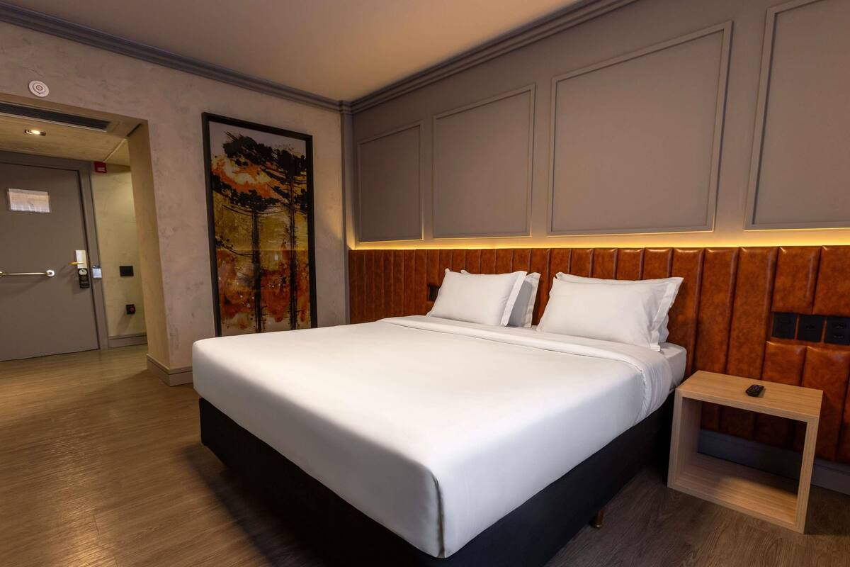 A modern hotel room with a large bed, a wooden headboard, a bedside table, and a painting on the wall.
