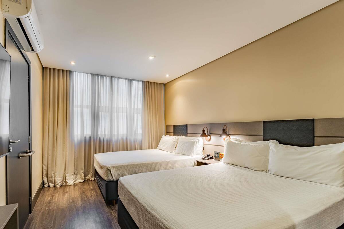 A hotel room with two beds, a desk, an air conditioning unit, and a window with curtains.