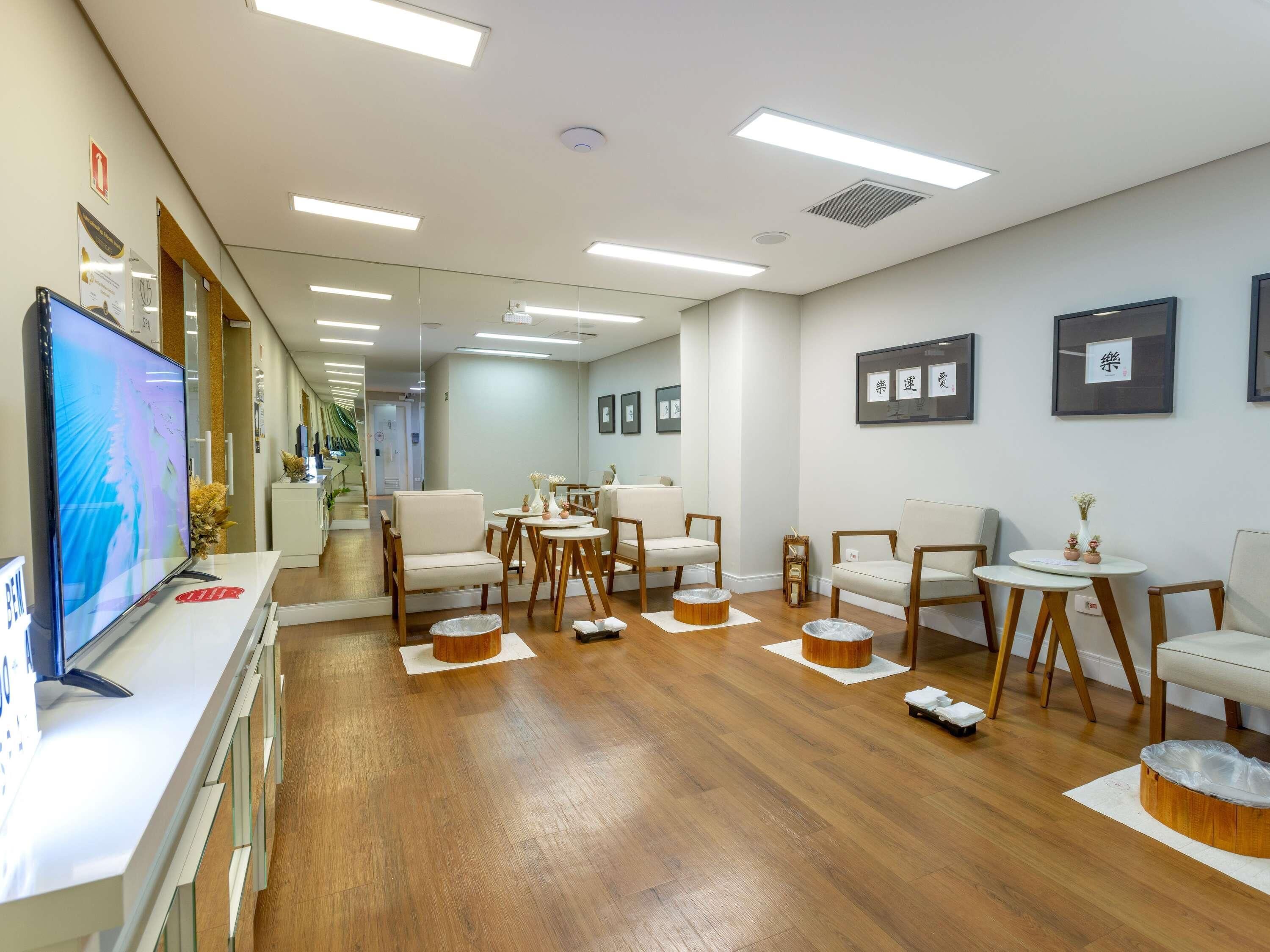 Un vestíbulo de hotel con suelo de madera, paredes blancas y un mostrador de recepción con televisión. Varias zonas de estar con sillas y mesas, y cuadros en las paredes.