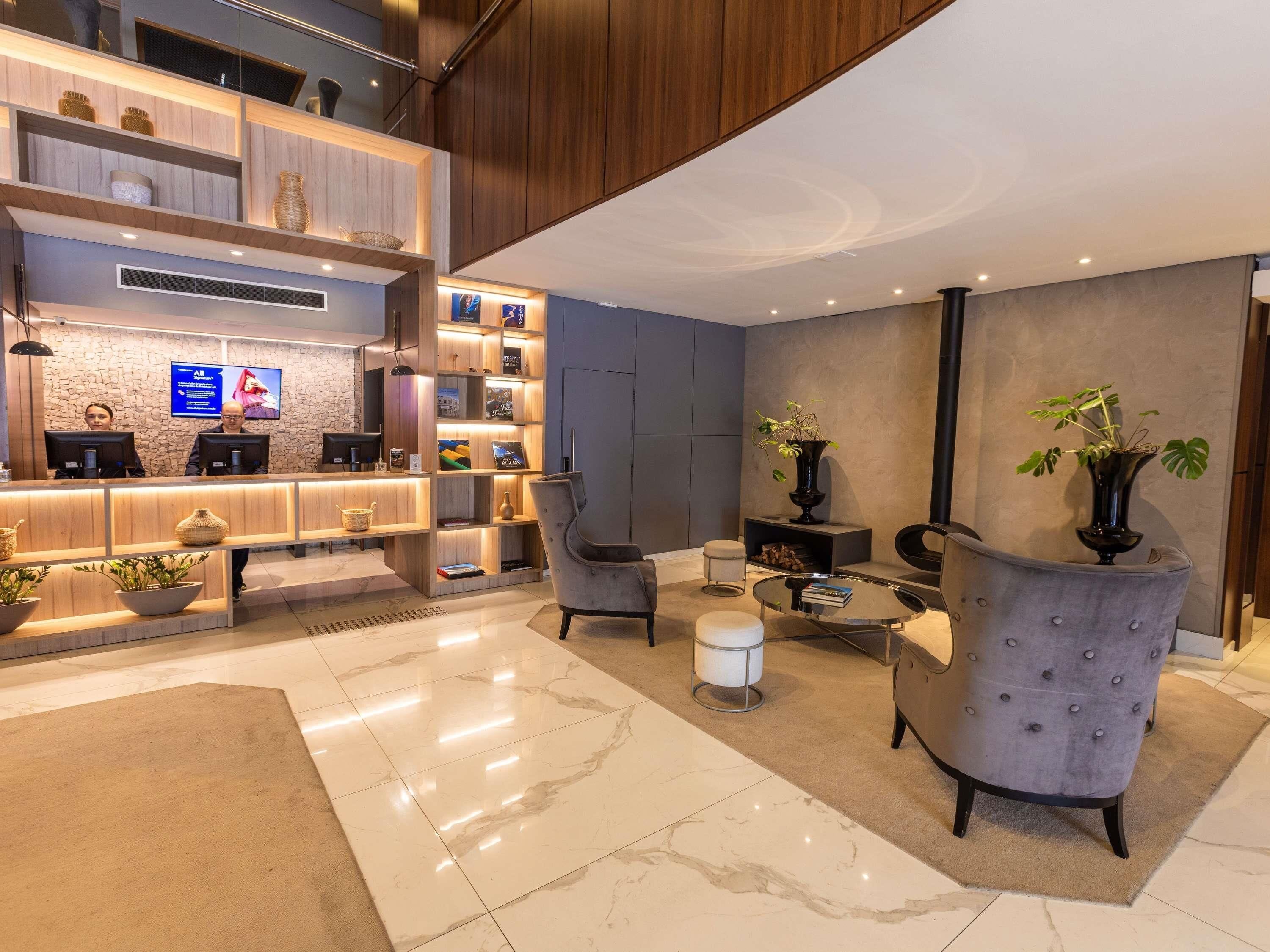 Un área de recepción moderna con un mostrador de madera, un televisor de pantalla plana y una pantalla mural. Hay dos zonas de estar con sillones y una pequeña mesa redonda.