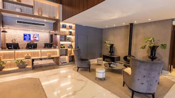 Un área de recepción moderna con un mostrador de madera, un televisor de pantalla plana y una pantalla mural. Hay dos zonas de estar con sillones y una pequeña mesa redonda.