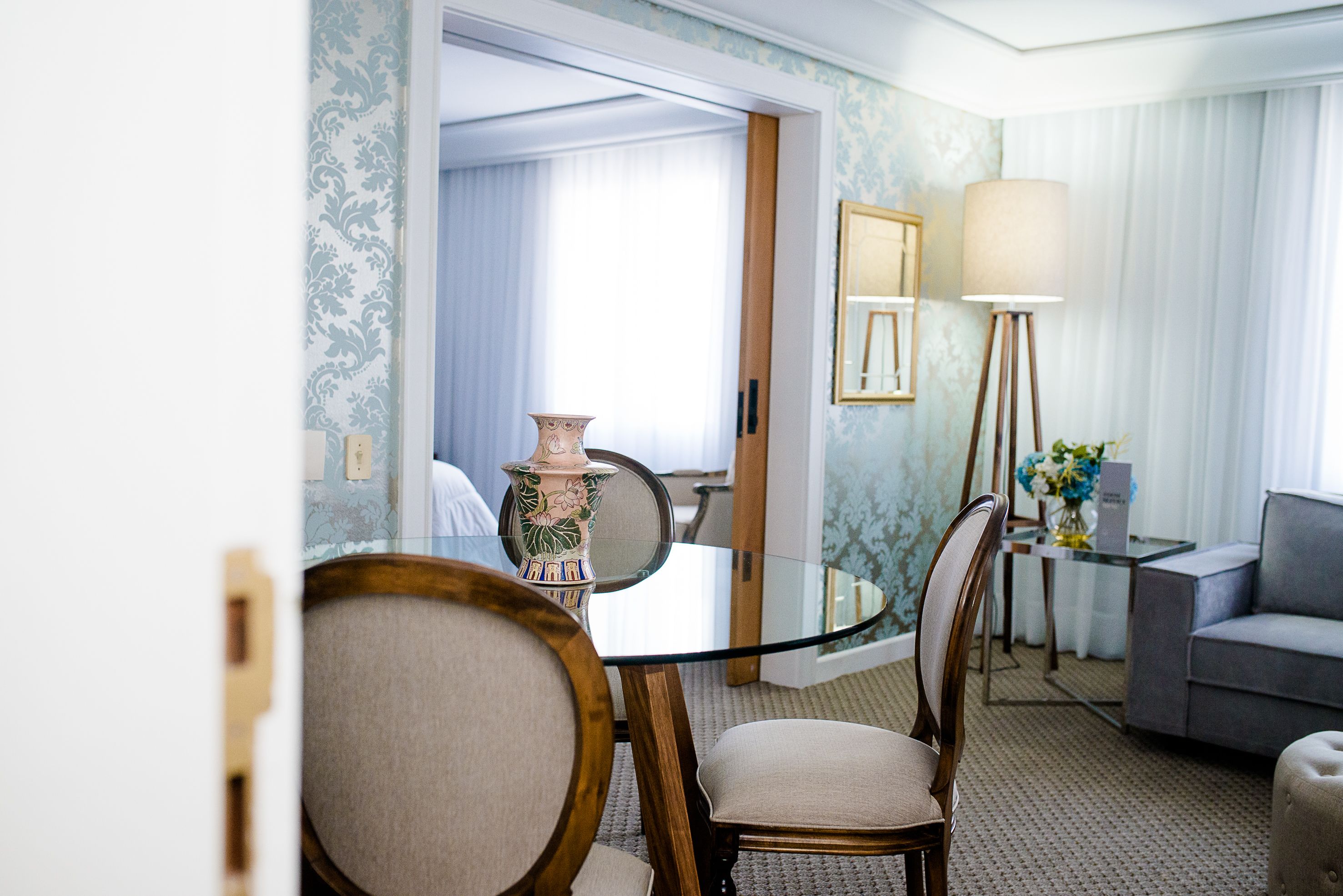 Une chambre d’hôtel comprenant une table à manger avec un plateau en verre, deux fauteuils rembourrés, un canapé gris, une lampe sur pied et un vase posé sur une table d’appoint.