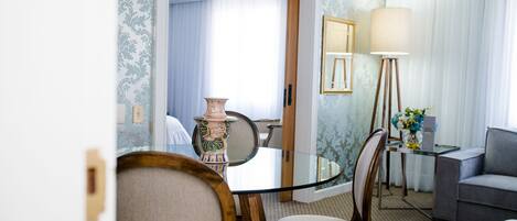 Une chambre d’hôtel comprenant une table à manger avec un plateau en verre, deux fauteuils rembourrés, un canapé gris, une lampe sur pied et un vase posé sur une table d’appoint.