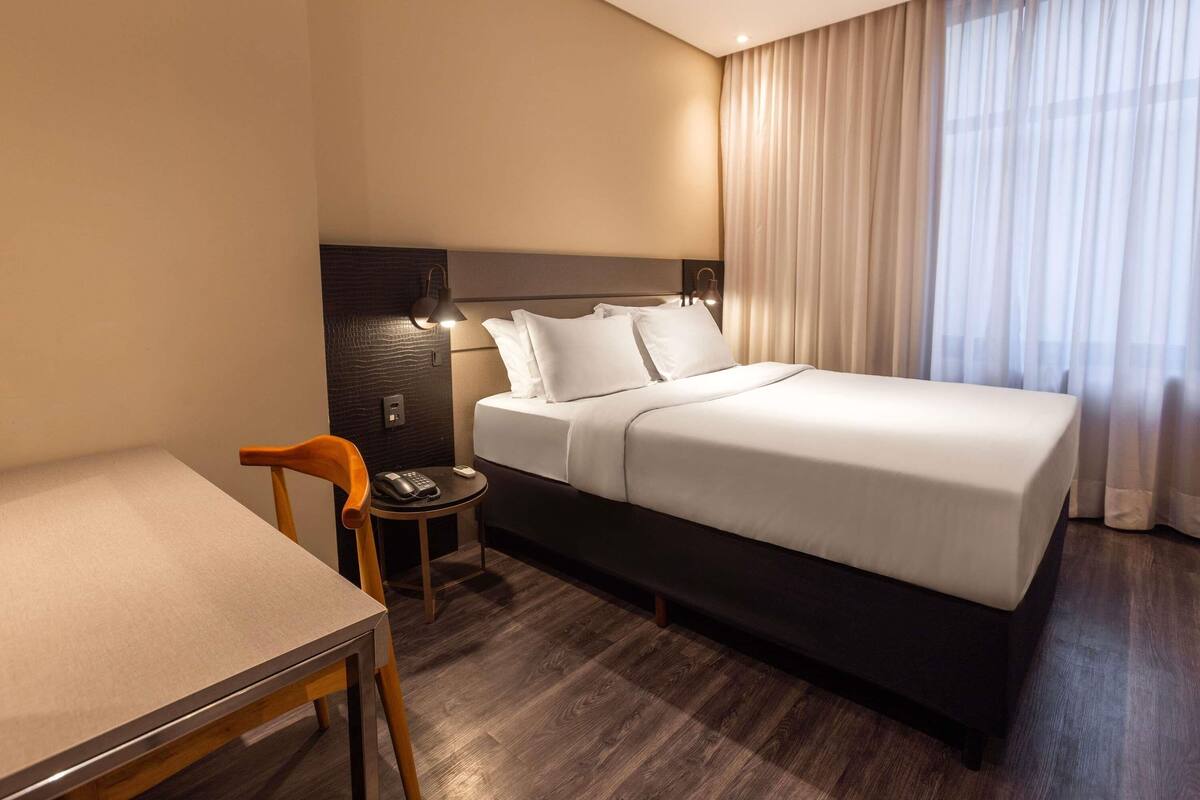 A hotel room with a bed, a desk, a chair, a telephone, and a lamp.