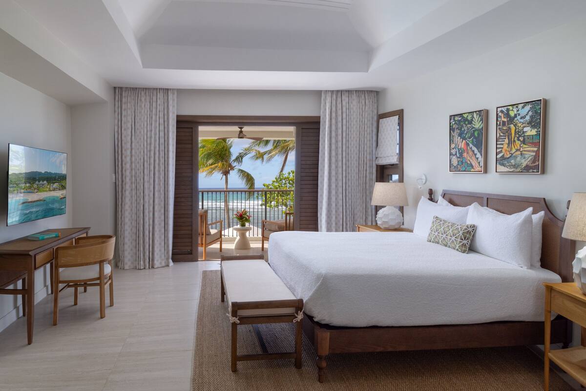 Une chambre d’hôtel équipée d’un lit, d’un bureau, de chaises, d’une télévision, d’œuvres d’art et d’un balcon offrant une vue sur les palmiers et l’océan.