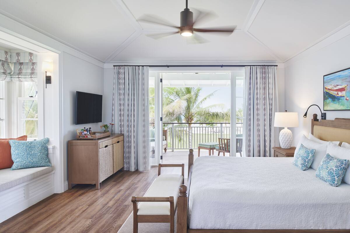 Une chambre d’hôtel comprenant un lit, un bureau, une télévision, un balcon avec des rideaux et un ventilateur de plafond.
