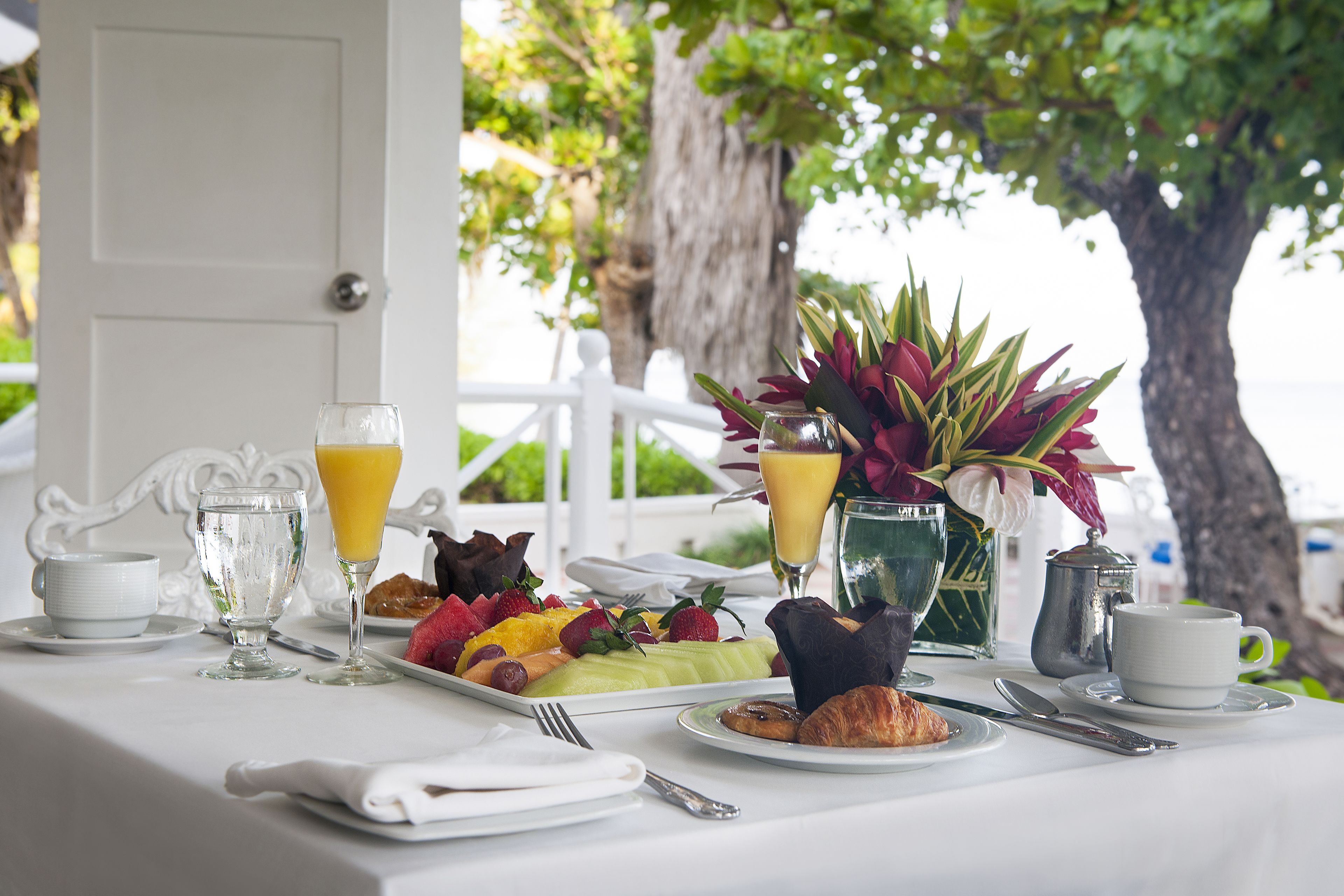 A breakfast table with a variety of food items, including fruits, pastries, and beverages, set against a backdrop of a white door and a tree.
