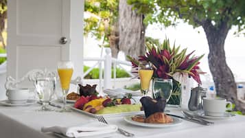 A breakfast table with a variety of food items, including fruits, pastries, and beverages, set against a backdrop of a white door and a tree.