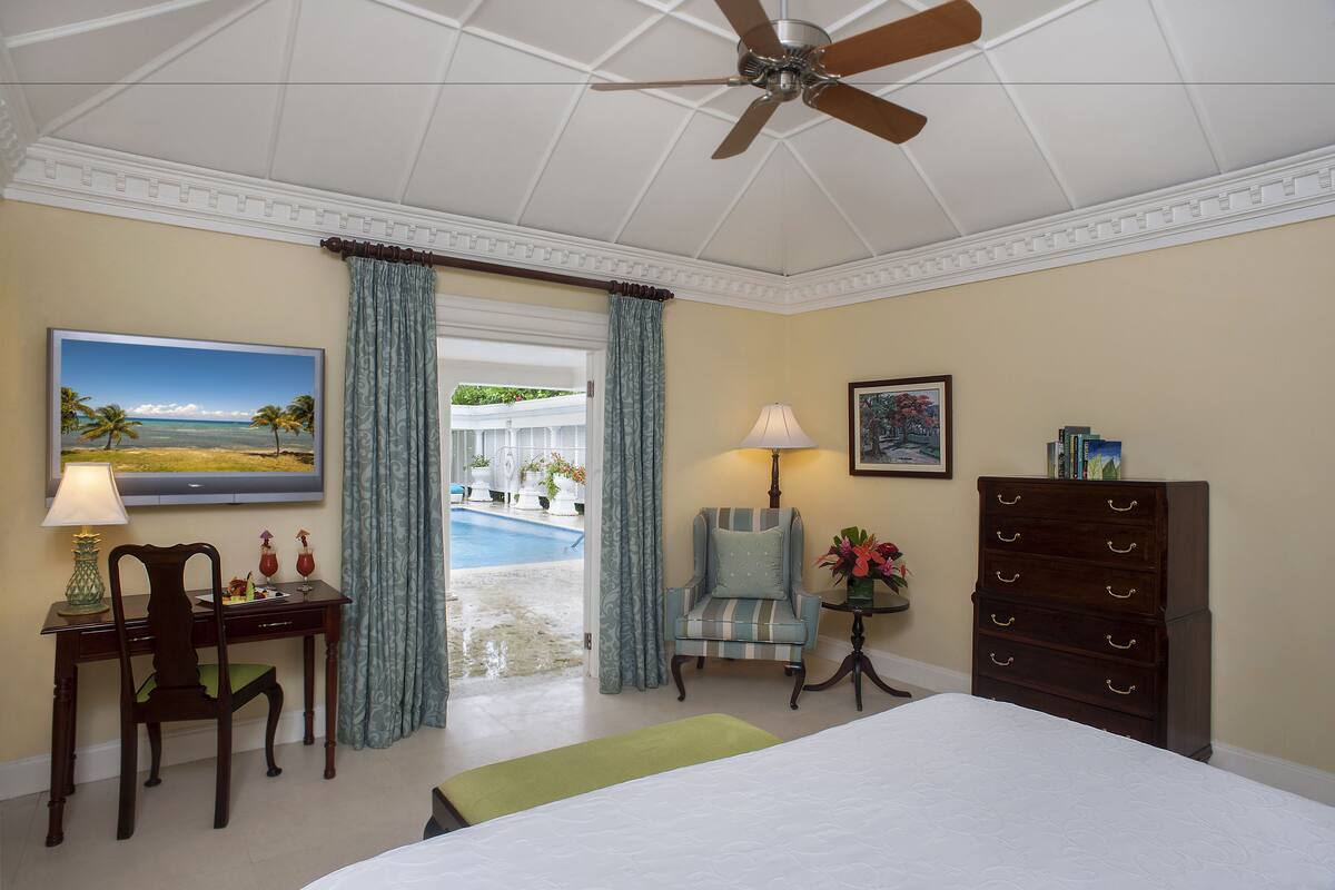 Une chambre d’hôtel comprenant un lit, un bureau avec une chaise, une commode, un ventilateur de plafond, un tableau, une lampe et une vue sur une piscine et des palmiers.
