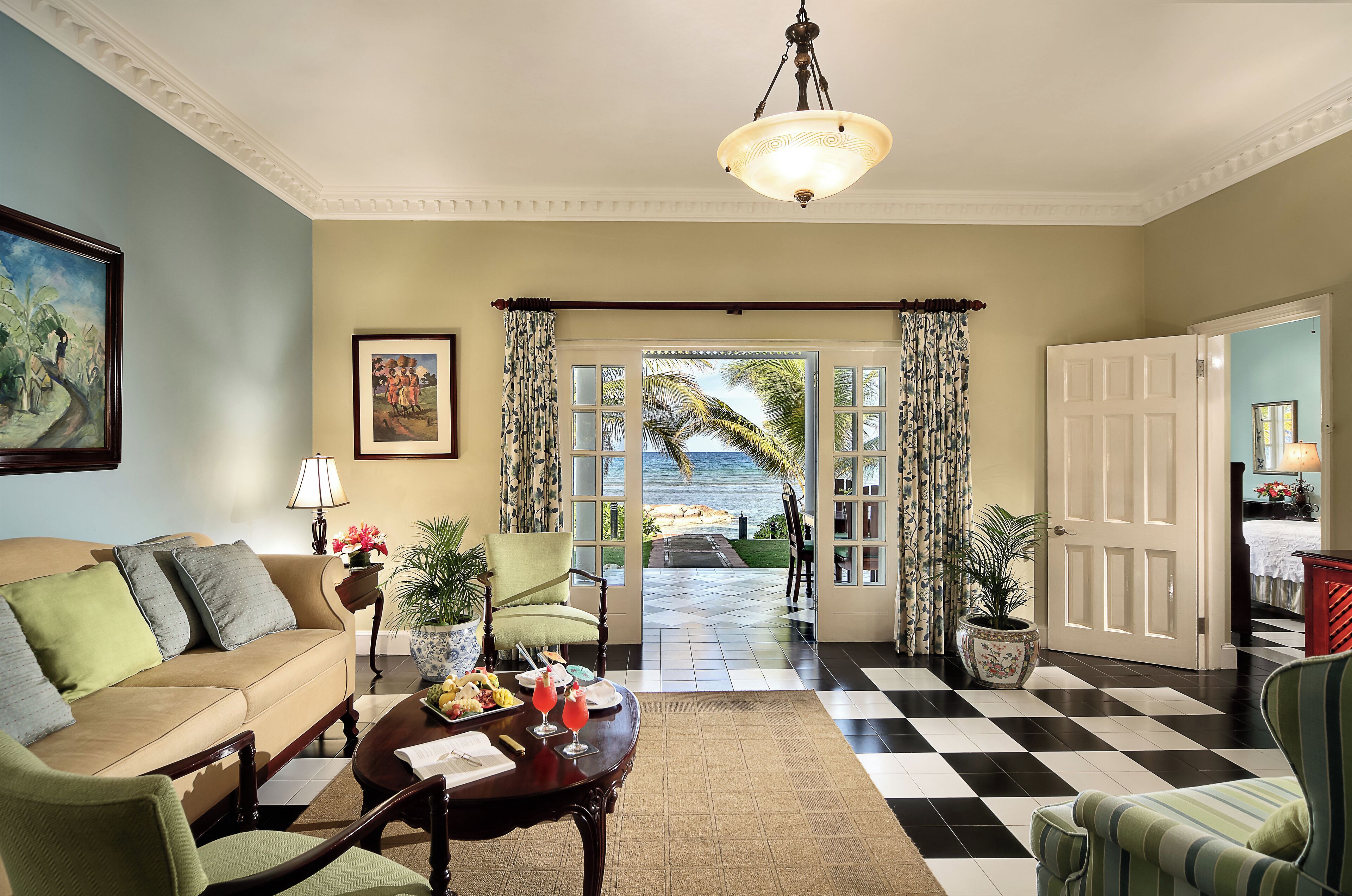 A living room with a sofa, armchairs, a coffee table, and a dining table set with food and drinks. There is a door leading to a balcony with a view of the ocean.