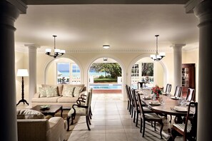A spacious dining area with a view of a pool and garden, featuring a long table set for dining, a sofa, and a lamp.