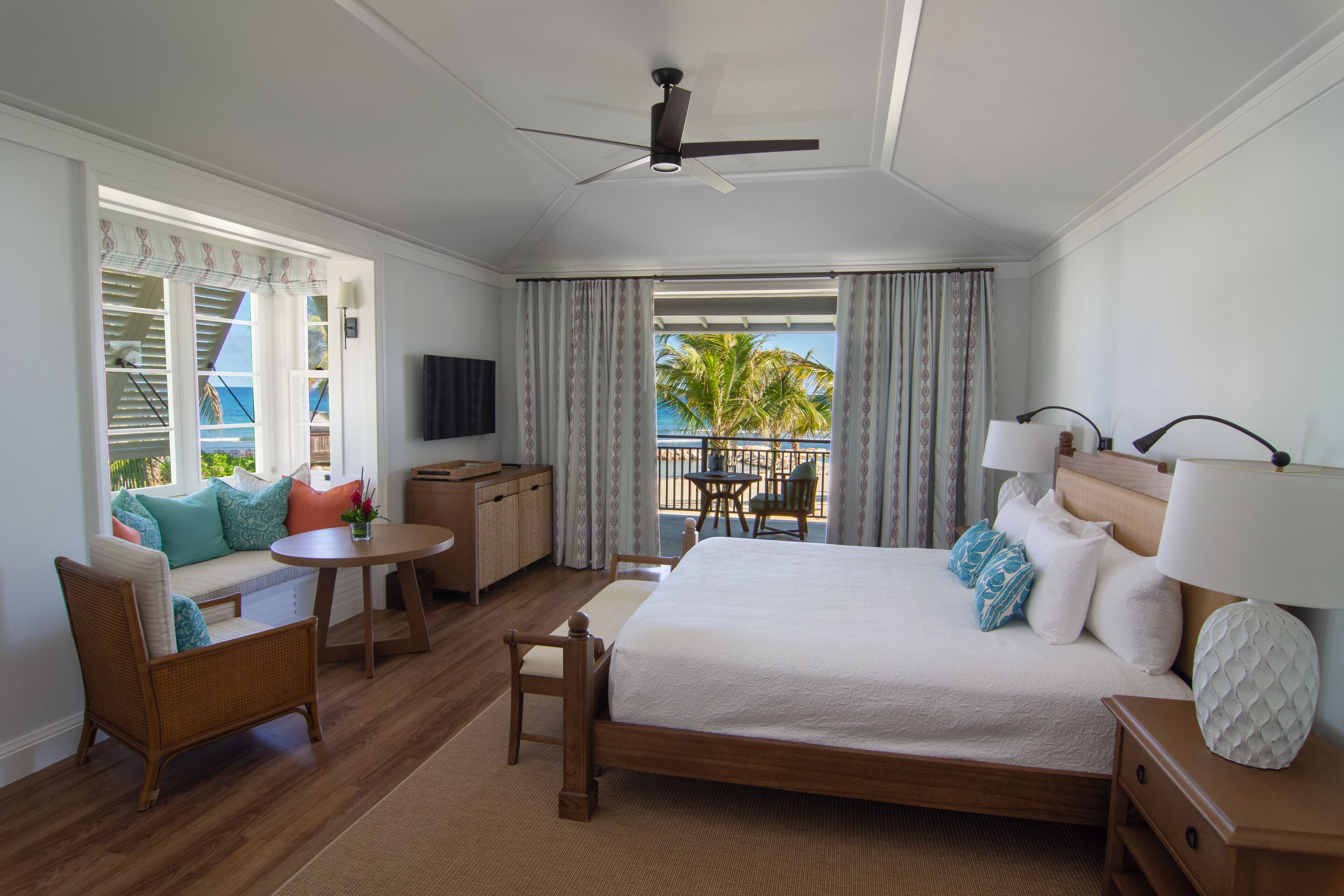 A hotel room with a bed, a desk, a chair, a small table, a ceiling fan, and a view of the ocean.