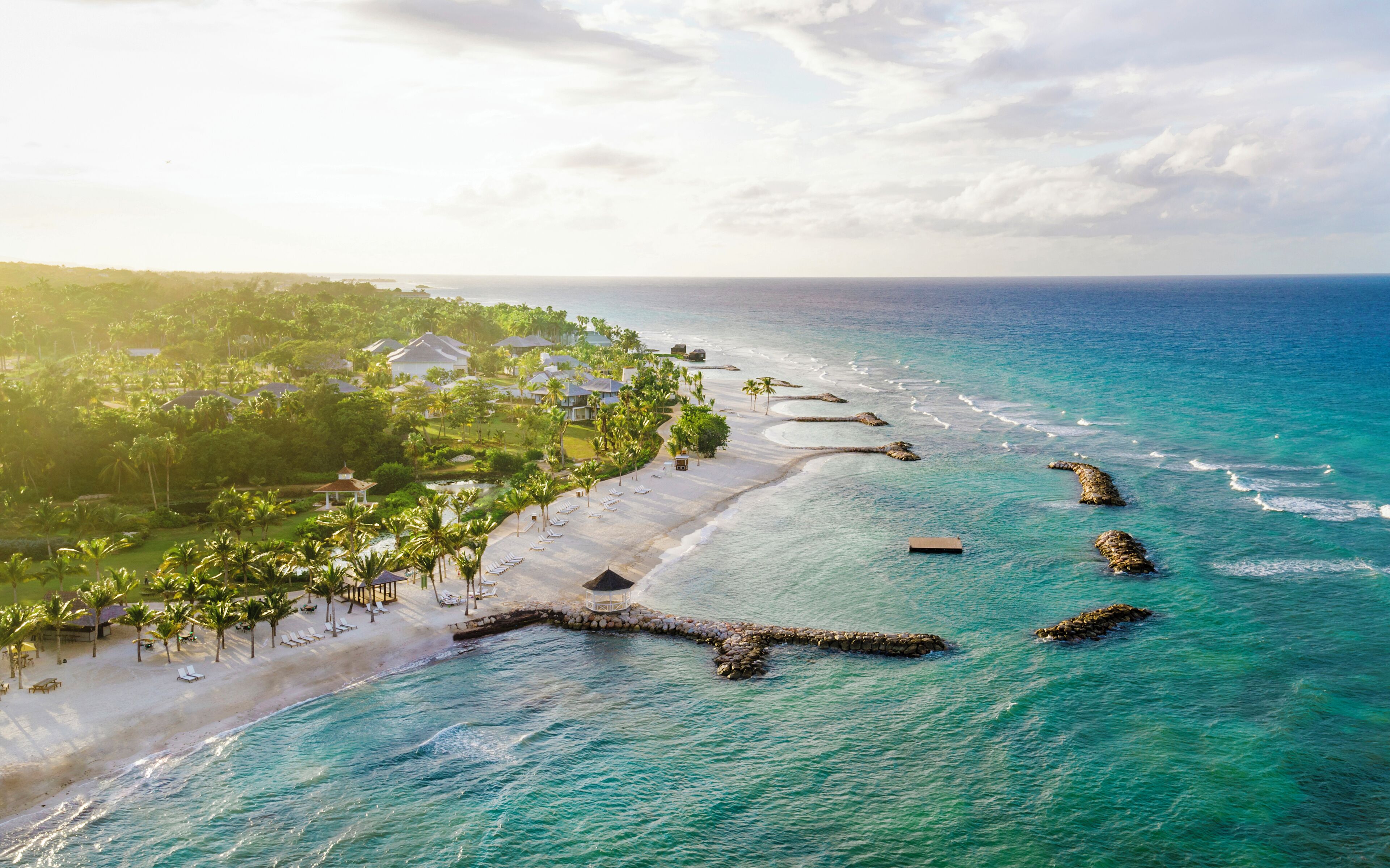 An aerial view of a beach resort with a long stretch of white sand, clear turquoise waters, and a line of palm trees.