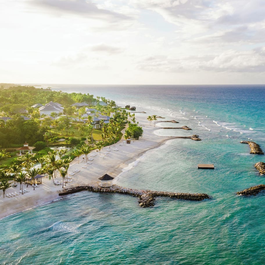 An aerial view of a beach resort with a long stretch of white sand, clear turquoise waters, and a line of palm trees.