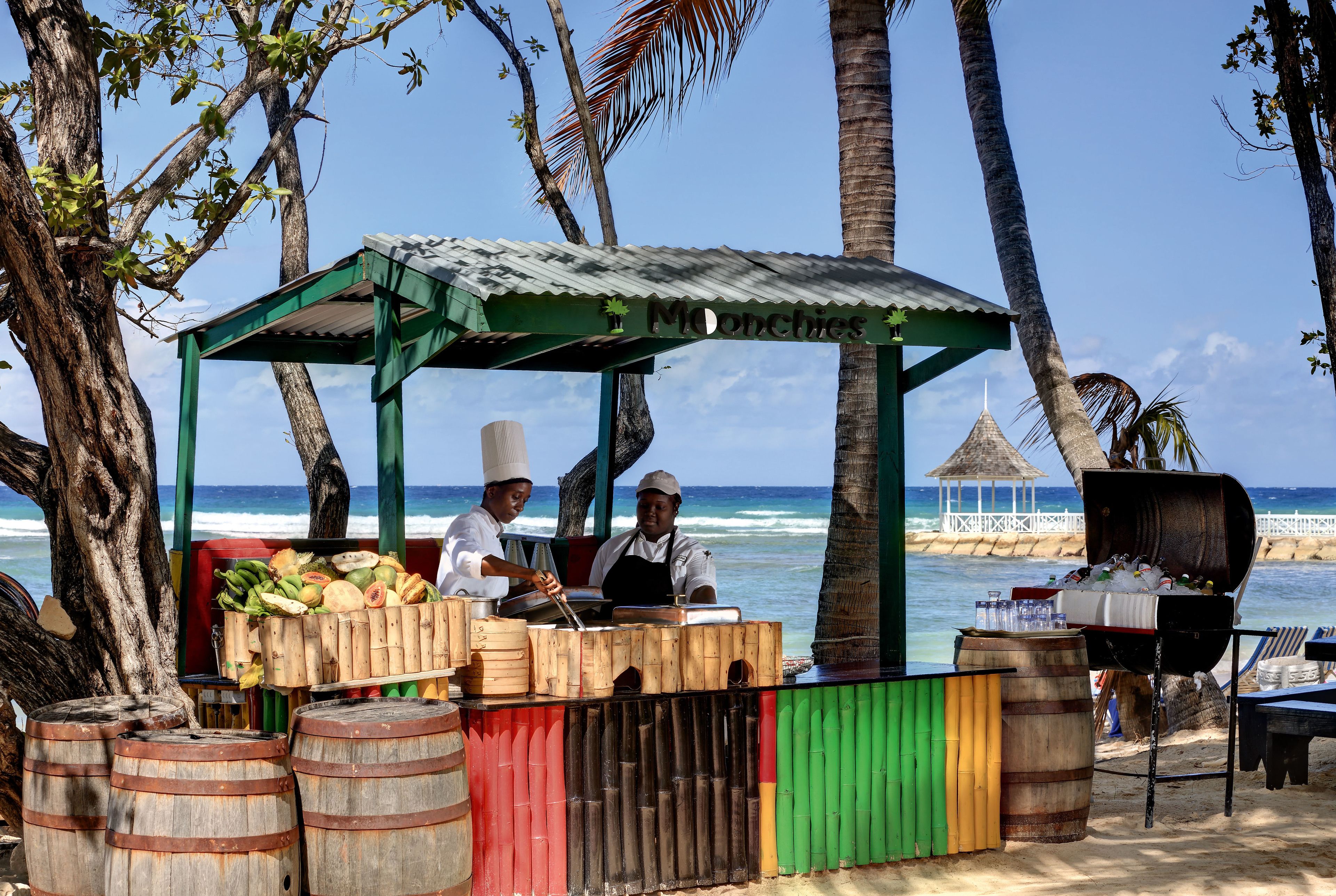 A beachside food stall with a green canopy, wooden barrels, and a view of the ocean.