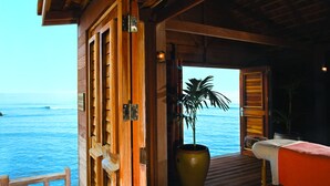 Habitación de madera con vista al océano, una planta grande en maceta y una cama con una colcha roja.