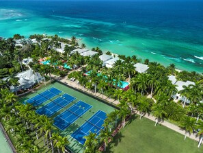An aerial view of a resort with a tennis court, palm trees, and a beach.