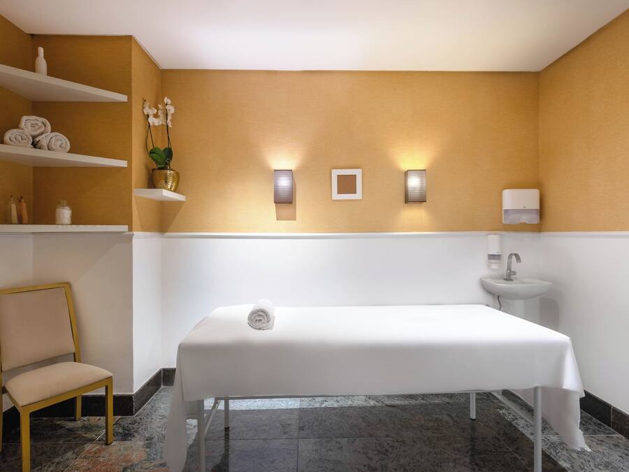 Um espaço de spa com mesa de massagem, uma cadeira, prateleiras com toalhas, pia e iluminação na parede.