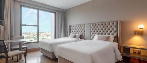 Habitación de hotel con dos camas, una zona de comedor y un amplio ventanal con vistas a la ciudad.