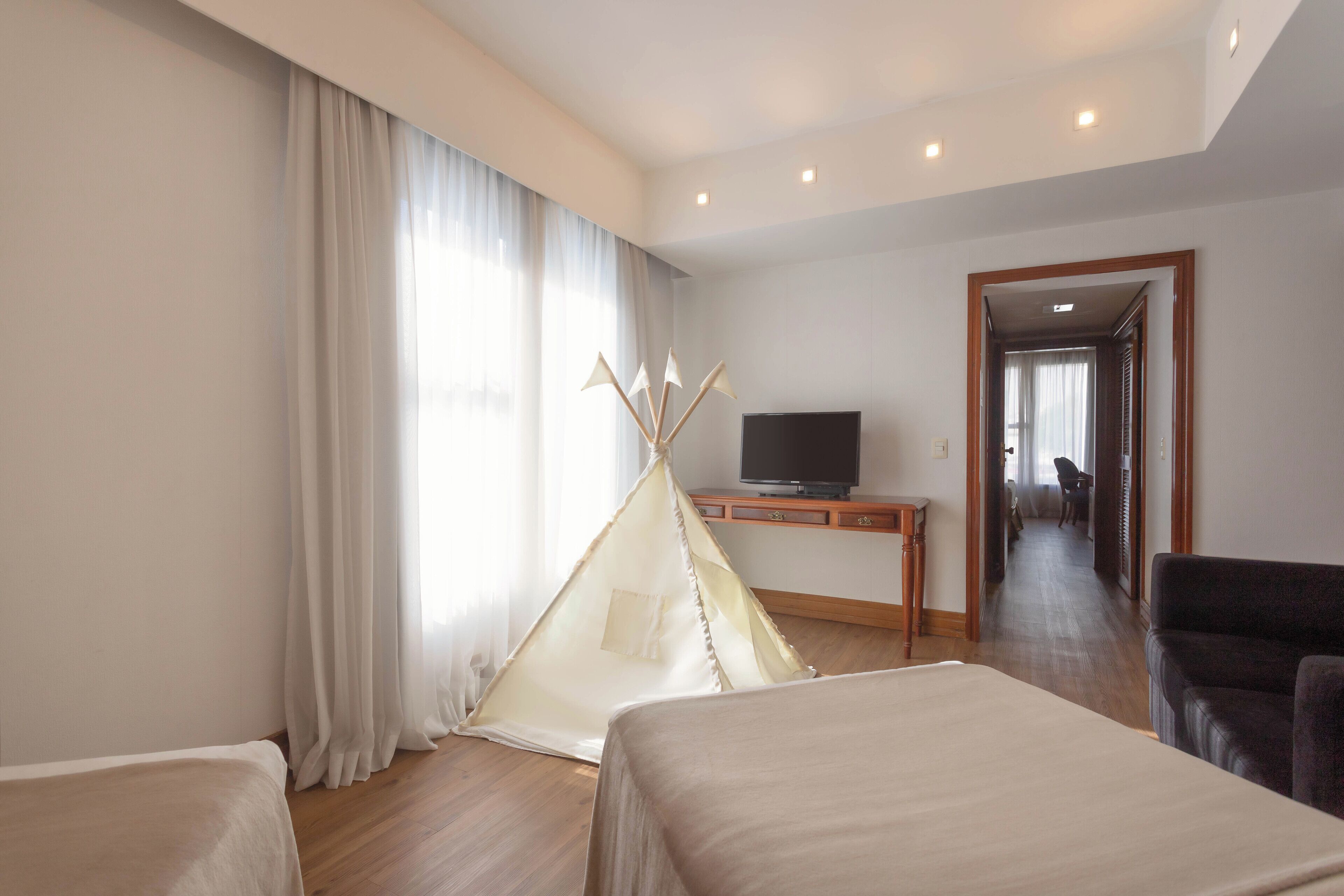 A hotel room with a bed, a TV on a wooden stand, a large window with curtains, and a hallway leading to another room.