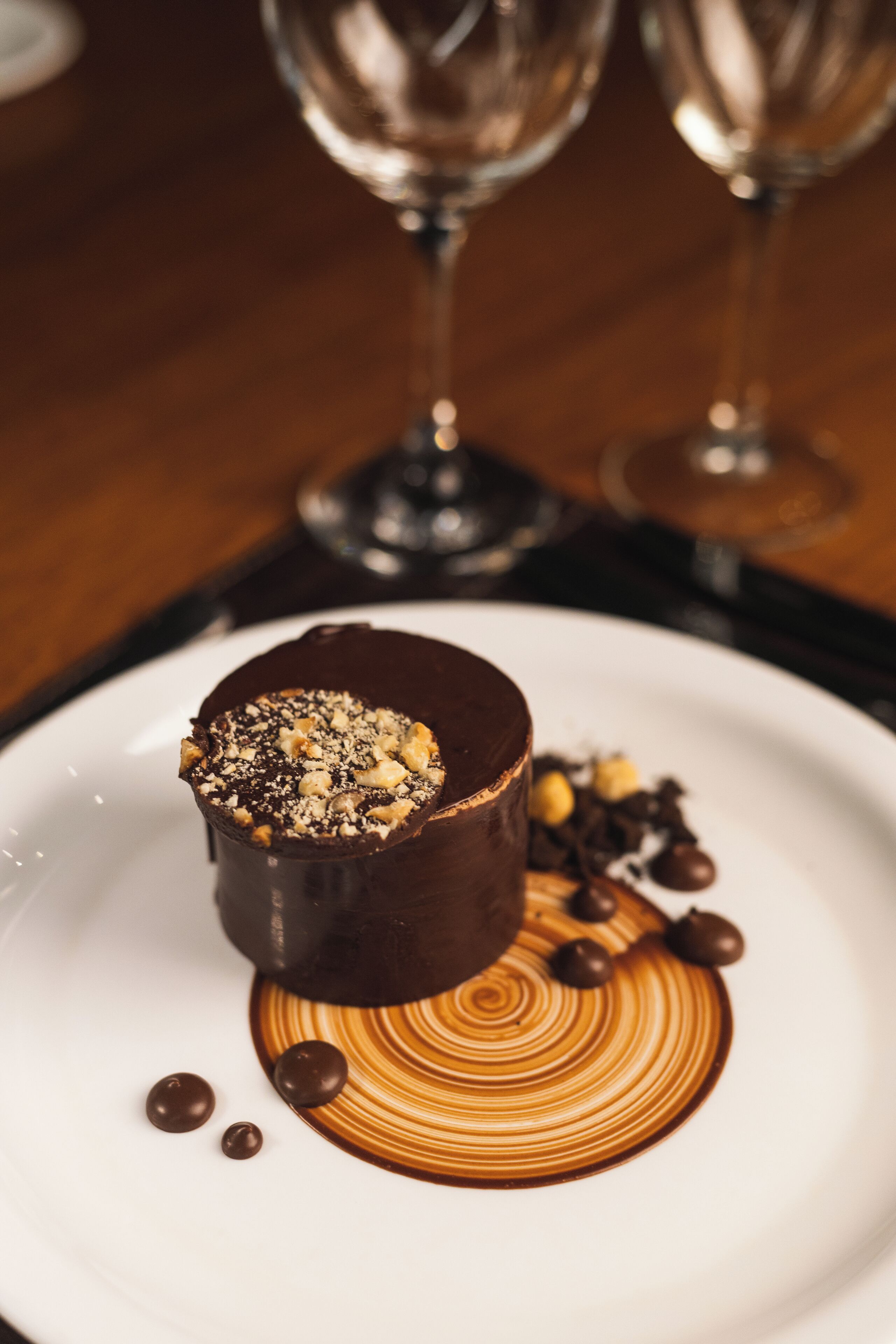 Sobremesa em prato branco com calda de chocolate e nozes, acompanhada por duas taças de vinho ao fundo.