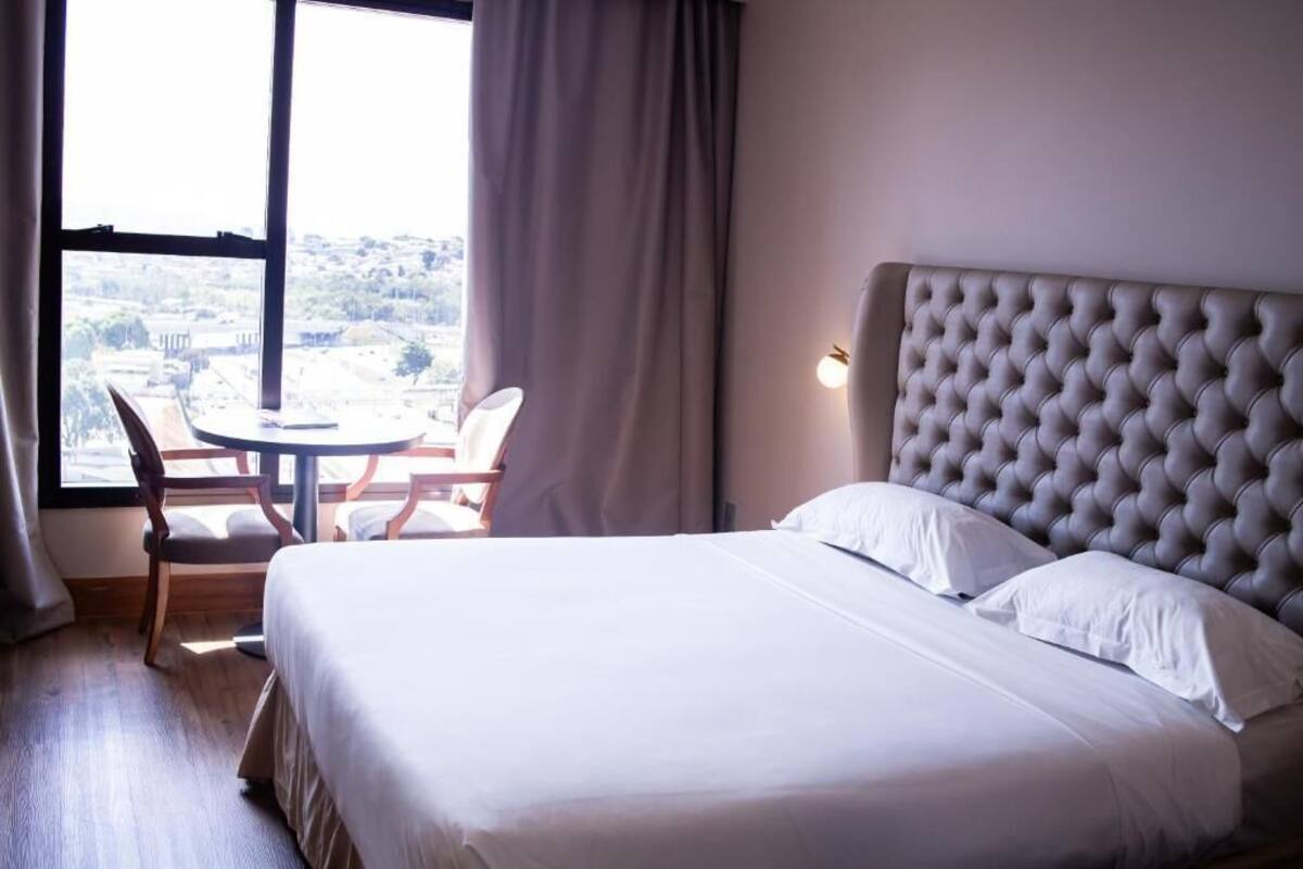 Een hotelkamer met een bed, een ronde tafel en stoelen, uitzicht door grote ramen en een gestoffeerd hoofdeinde.