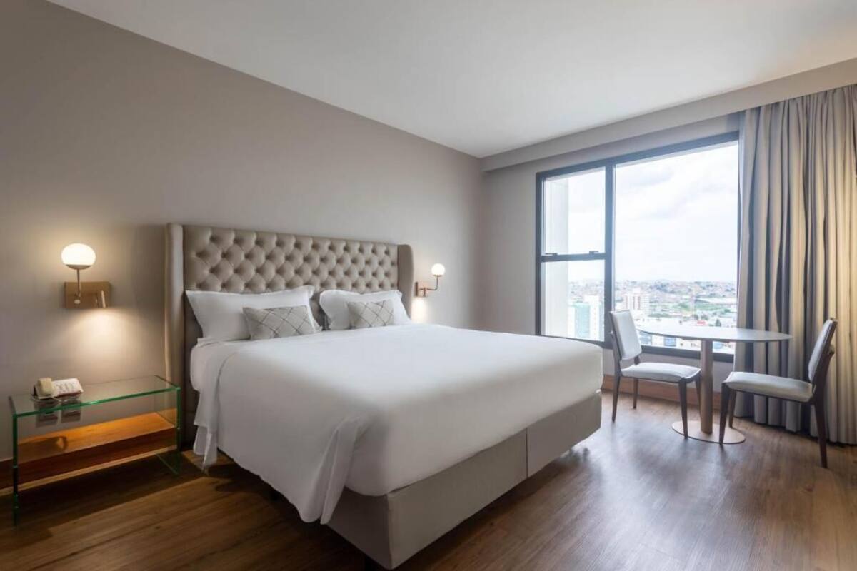 Een moderne hotelkamer met een groot bed, een glazen bijzettafeltje, een kleine tafel met stoelen en een stadszicht door grote ramen.