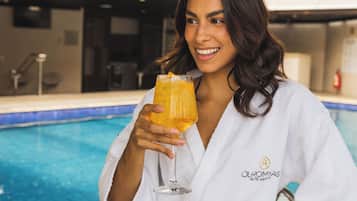 A person in a white robe holding a glass of orange beverage by a poolside.