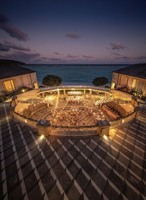 An outdoor dining area with tables and chairs arranged in a circular pattern, surrounded by a stone wall and a railing, overlooking a body of water. - Mandarin Oriental, Canouan (Canouan Island)