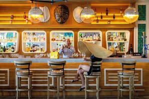 Un bar con taburetes de madera, una mujer con sombrero de paja y estantes llenos de botellas de licor.
