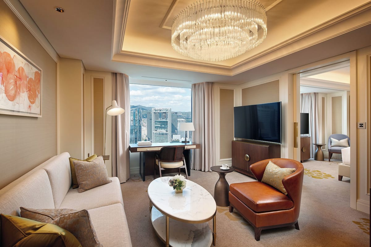 A hotel room with a large window, a chandelier, a flat-screen TV, a sofa, a coffee table, and a desk with a chair.