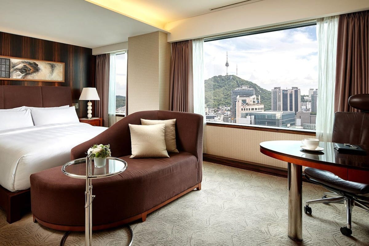 A hotel room with a bed, a brown armchair, a small table with a vase, a desk with a chair, and a large window with a city view.