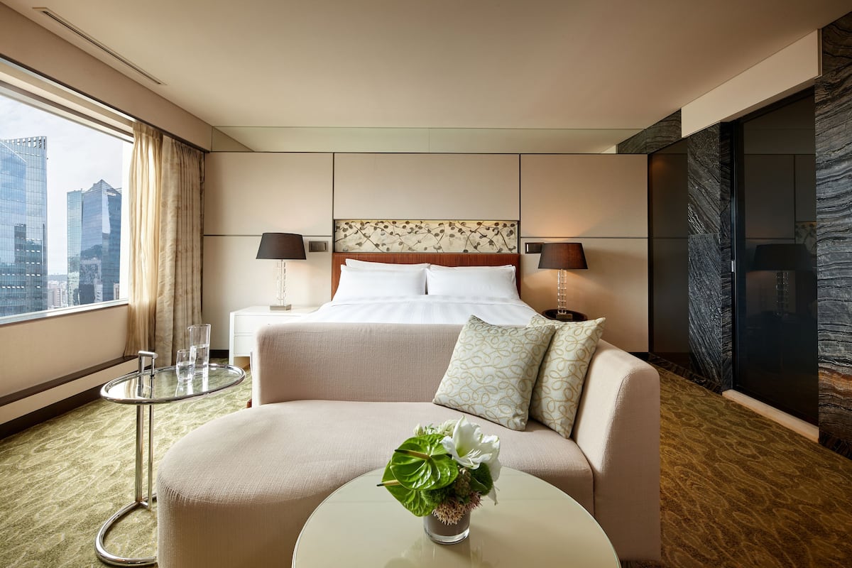 A hotel room with a bed, a sofa, a small table with a vase of flowers, a nightstand with lamps, and a view of the cityscape through large windows.