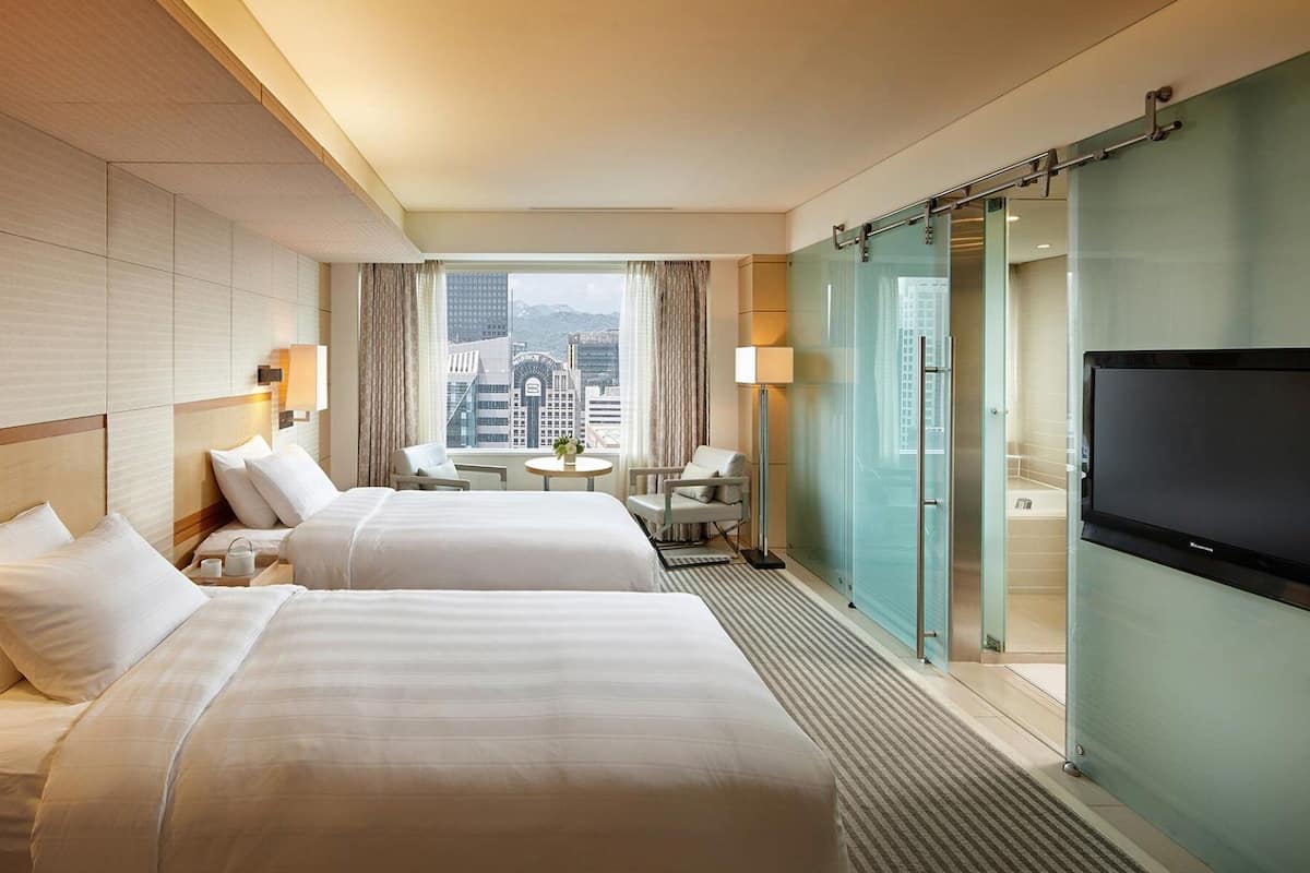 A hotel room with two beds, a TV, a small table with a chair, and a view of the city.