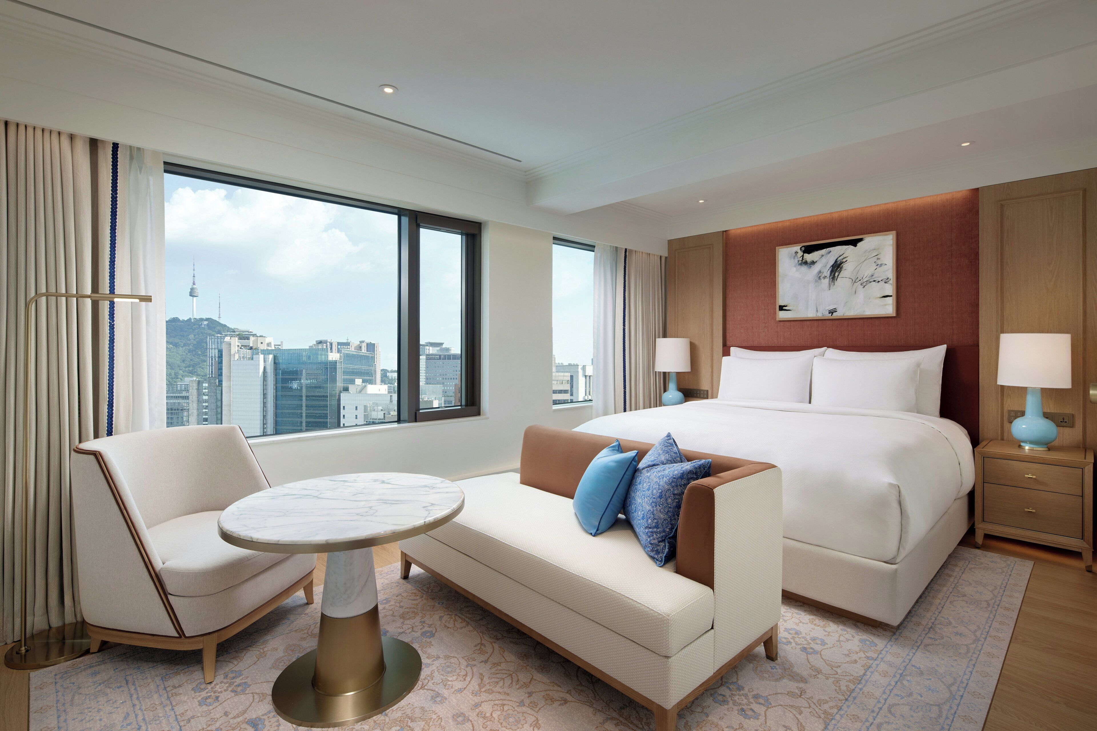 A hotel room with a bed, a sofa, a small table, and a view of the cityscape.