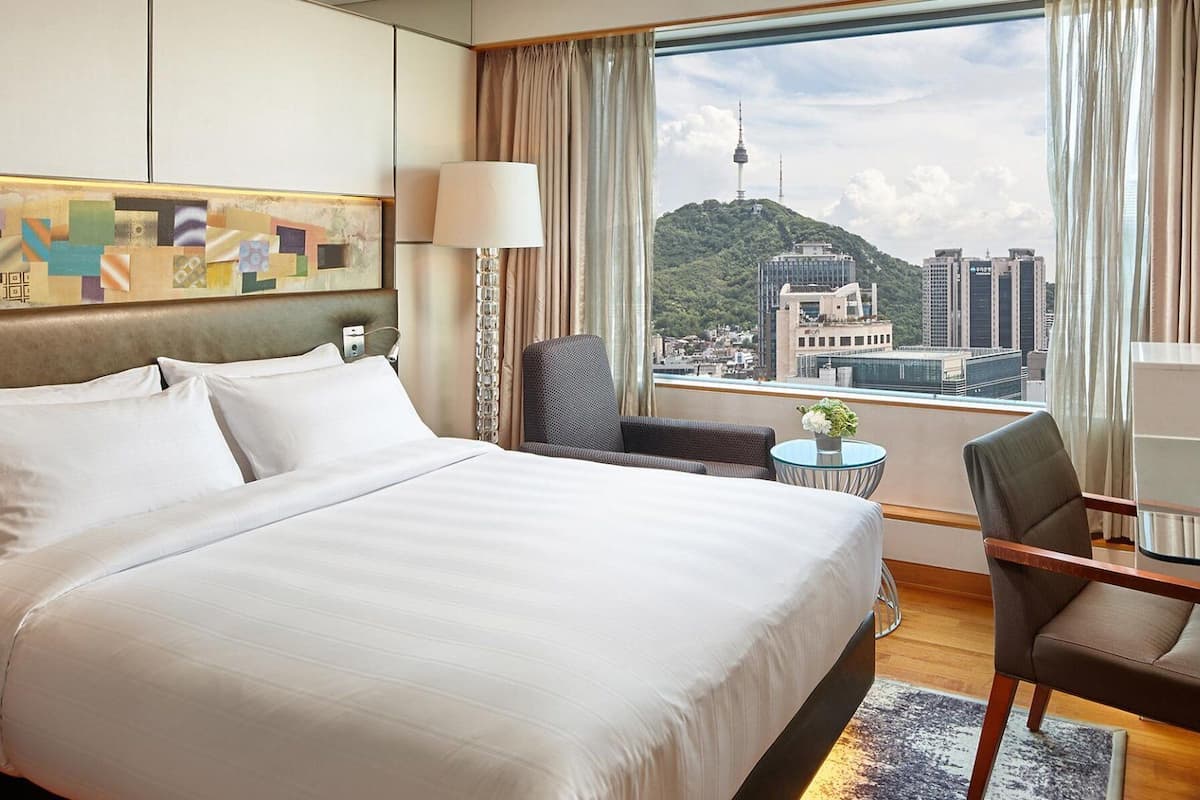 A hotel room with a large bed, a desk with a chair, a small table with a vase, and a view of a cityscape with a tower.